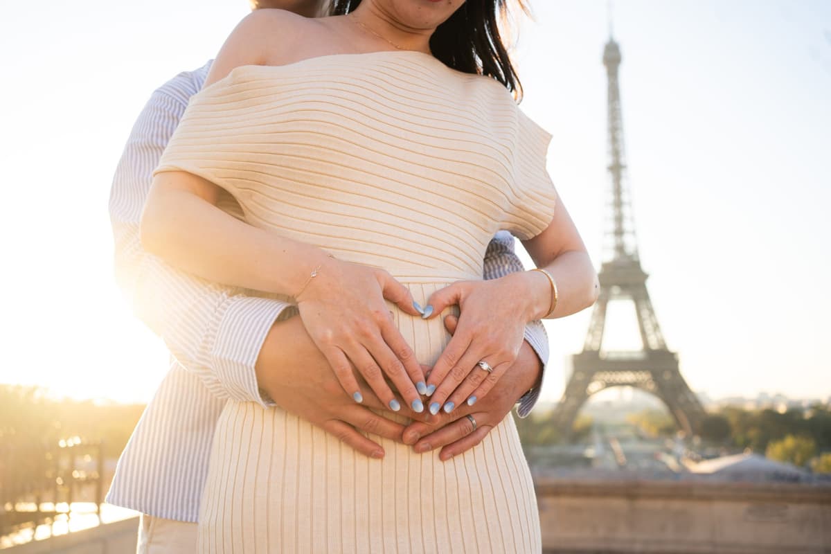 Expecting couple forming a heart shape over a baby bump during a maternity photoshoot in Paris with the Eiffel Tower in the background.