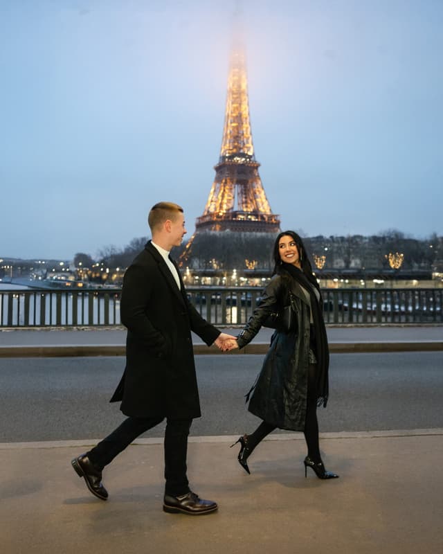 Couple walking hand-in-hand along the street at dusk with the Eiffel Tower glowing behind them during a Paris photo session.