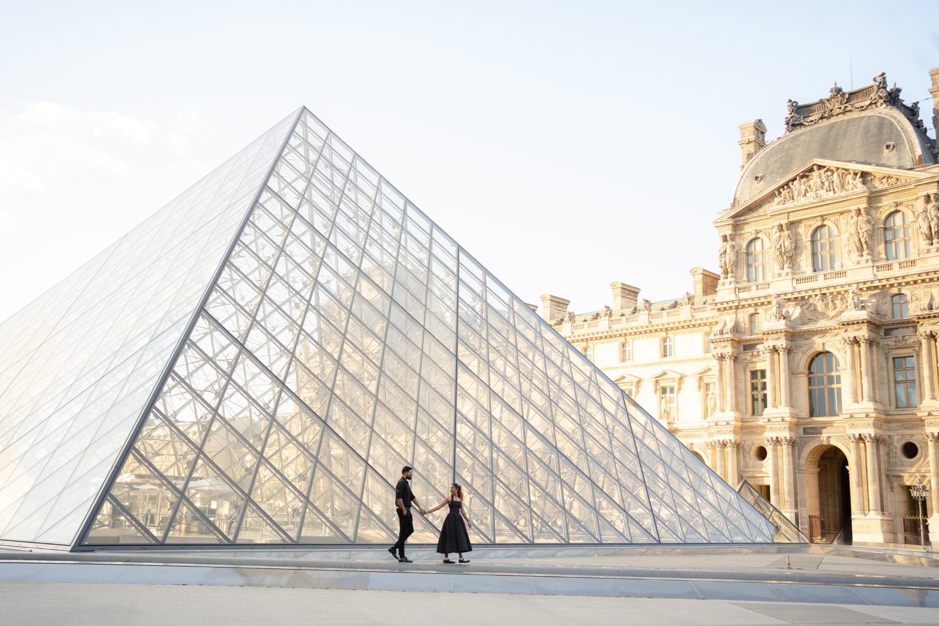 Couple holding hands while walking in front of the glass Louvre Pyramid during a romantic photo session in Paris.