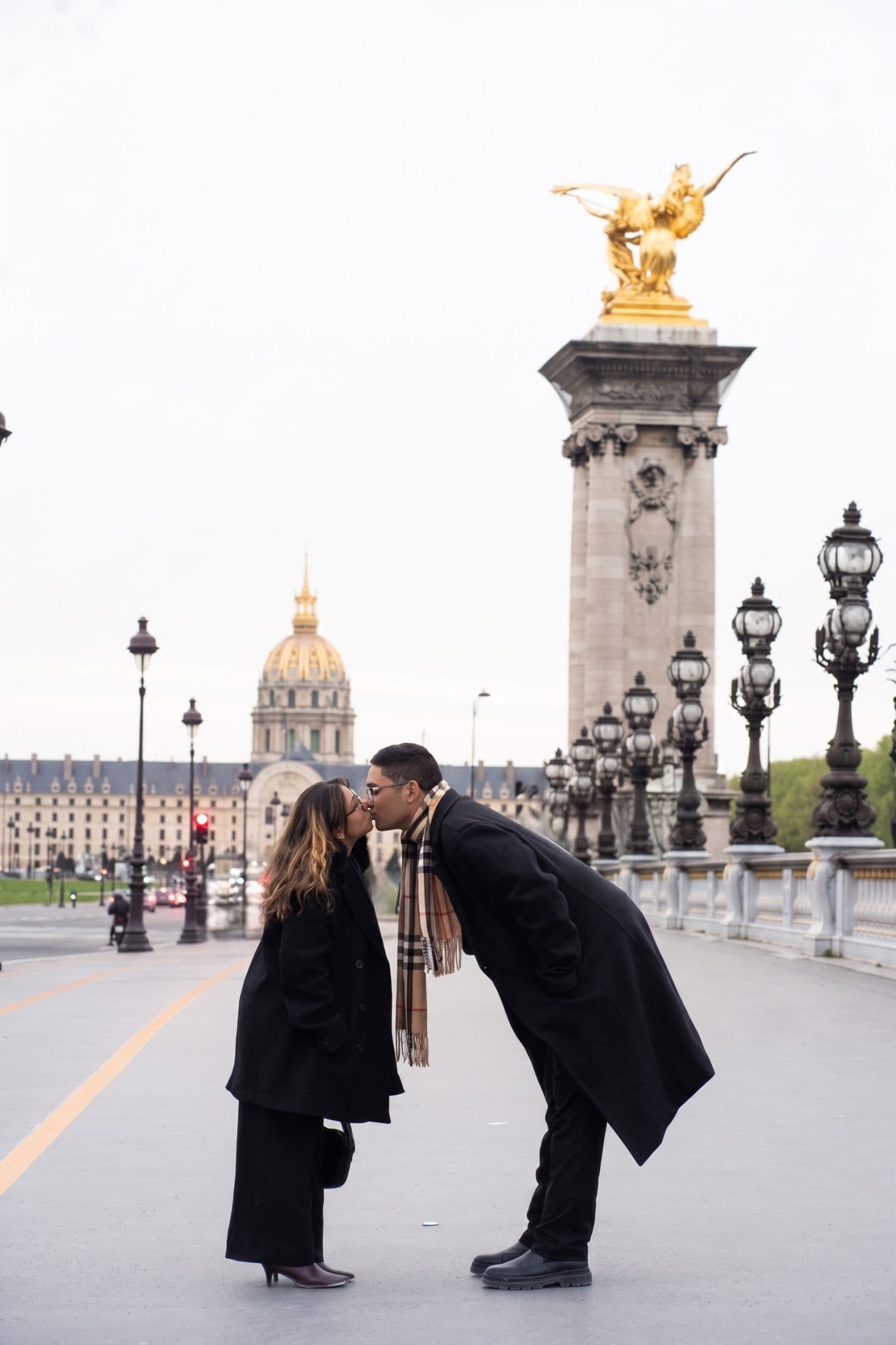 Couple leaning in for a kiss on Pont Alexandre III bridge with Les Invalides in the background during a Paris photoshoot.